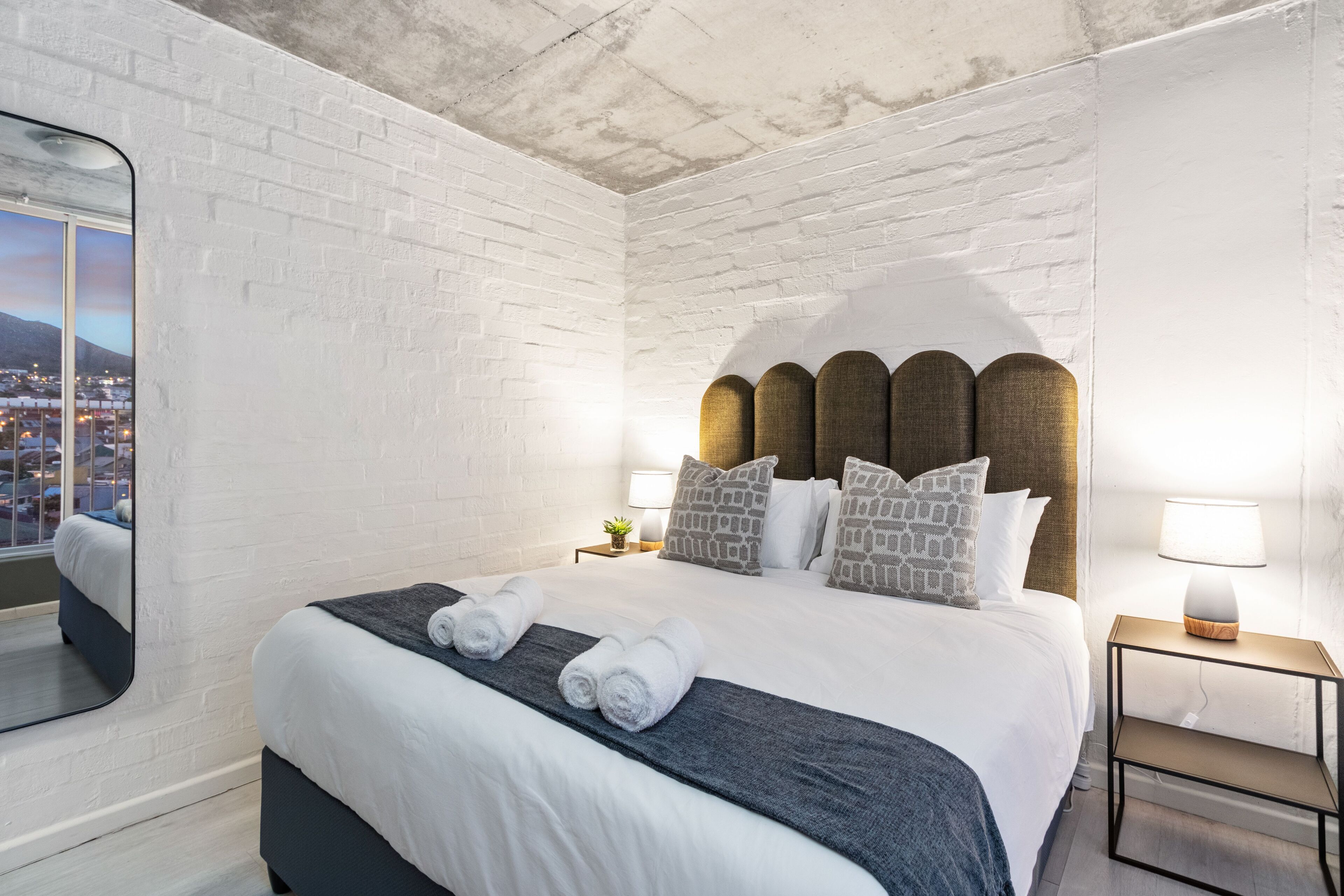 Quarto de hotel moderno com parede de tijolos brancos, uma cama grande com cabeceira original, mesa de cabeceira com abajur e vista para a paisagem urbana pela janela.