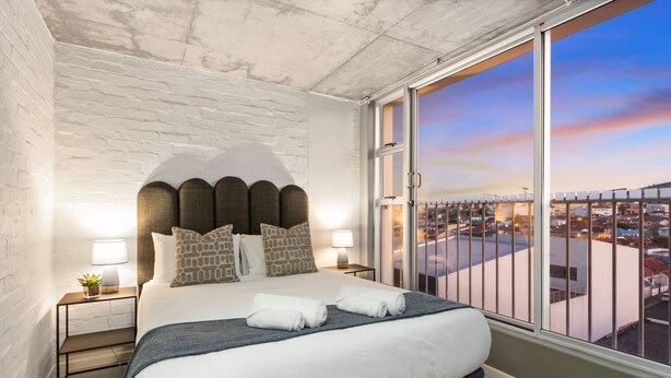 A bedroom with a large bed, a view of the city through a large window, and a brick wall.