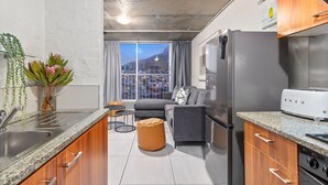 A modern kitchen with granite countertops, a stainless steel refrigerator, and a built-in oven. There is a dining area with a gray sofa and a small round table. A large window offers a view of a cityscape and a mountain.