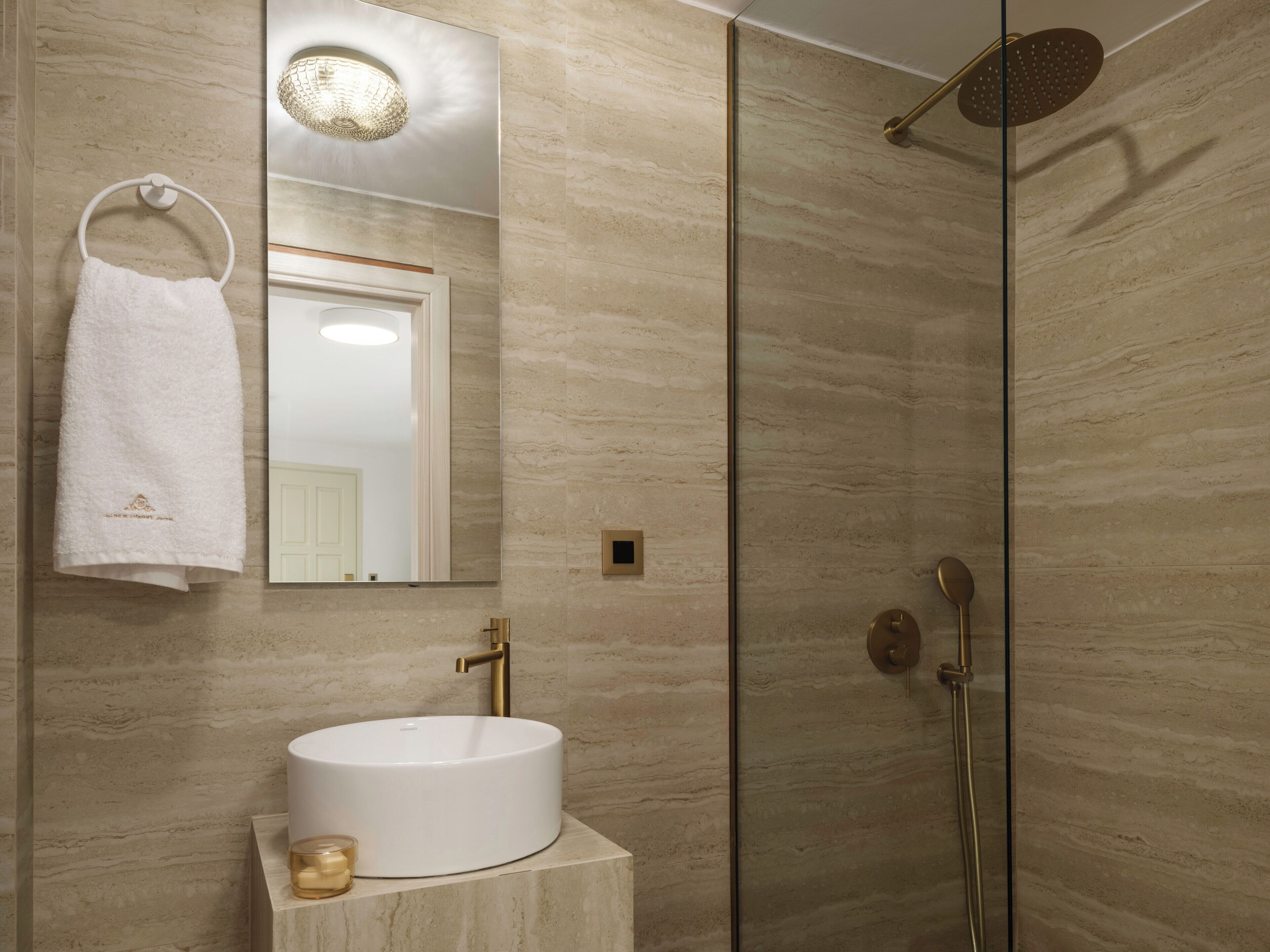 A modern bathroom with a round white sink, a towel rack with a white towel, a glass-enclosed shower, and a mirror with a textured light fixture.