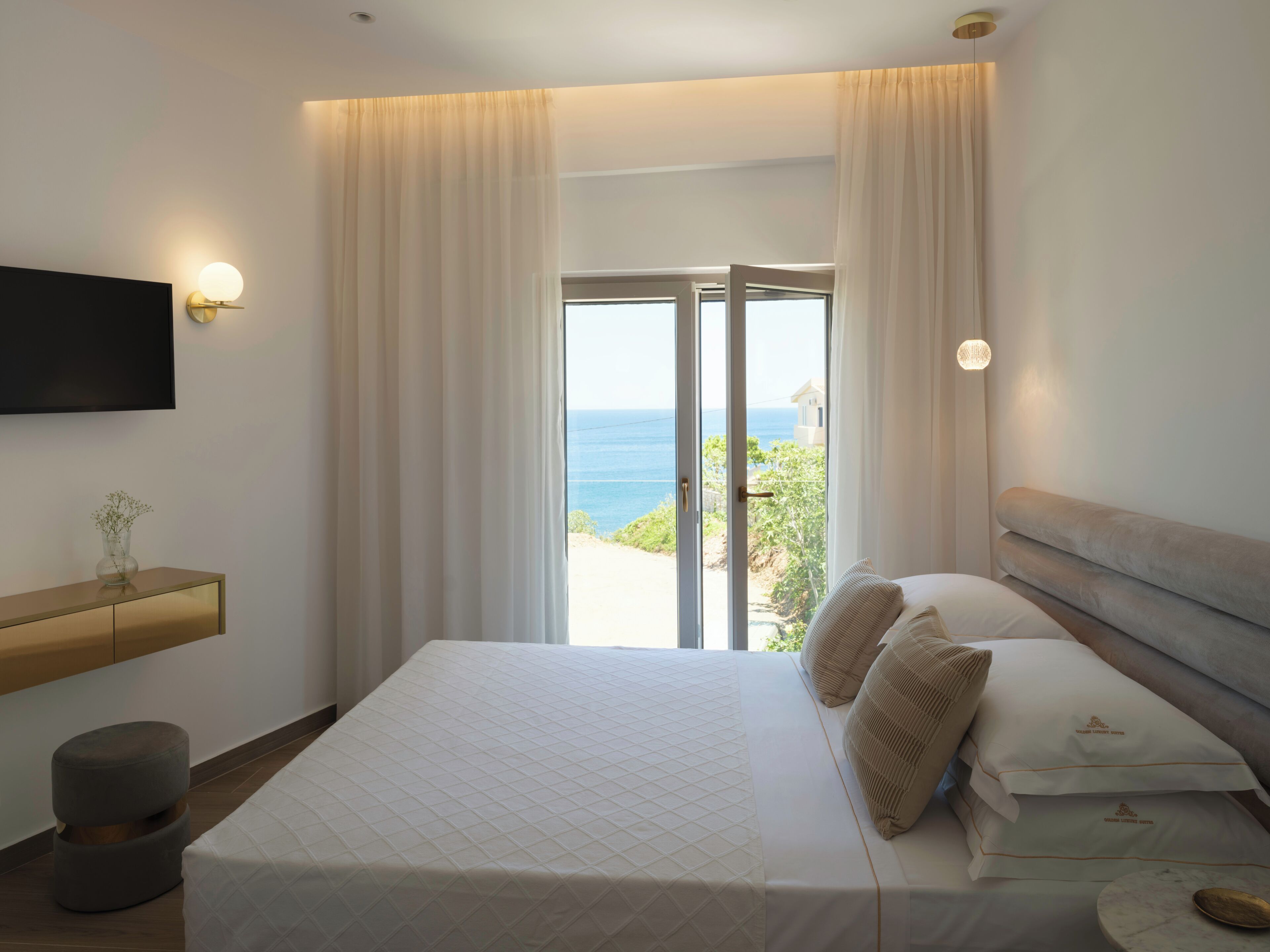 A bedroom with a bed, a nightstand, a TV, and a view of the sea through the window.