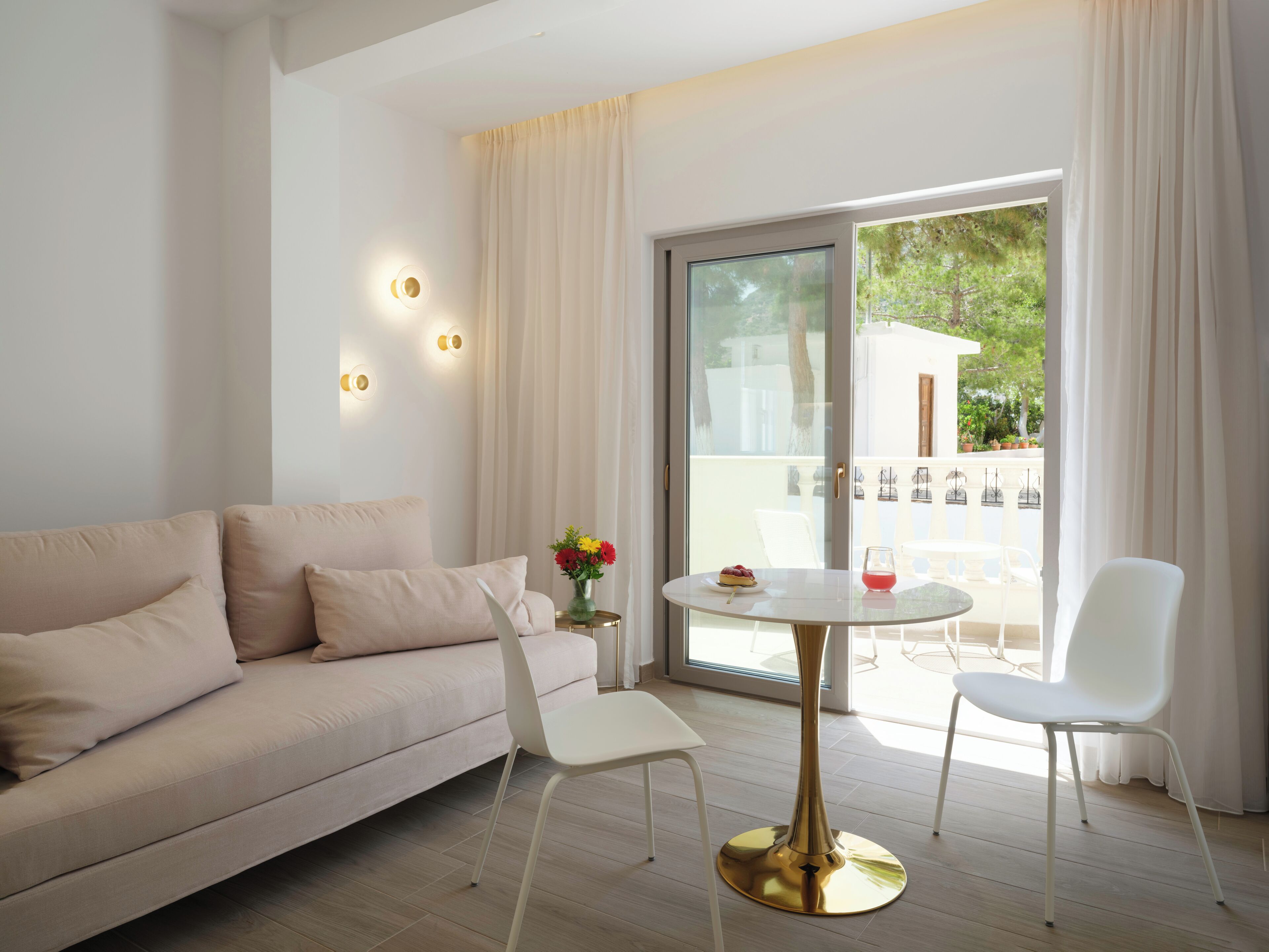 Una sala moderna con un sofá beige, una mesa redonda blanca y una silla blanca. Hay una puerta de vidrio que da a un espacio exterior con vistas a zonas verdes.