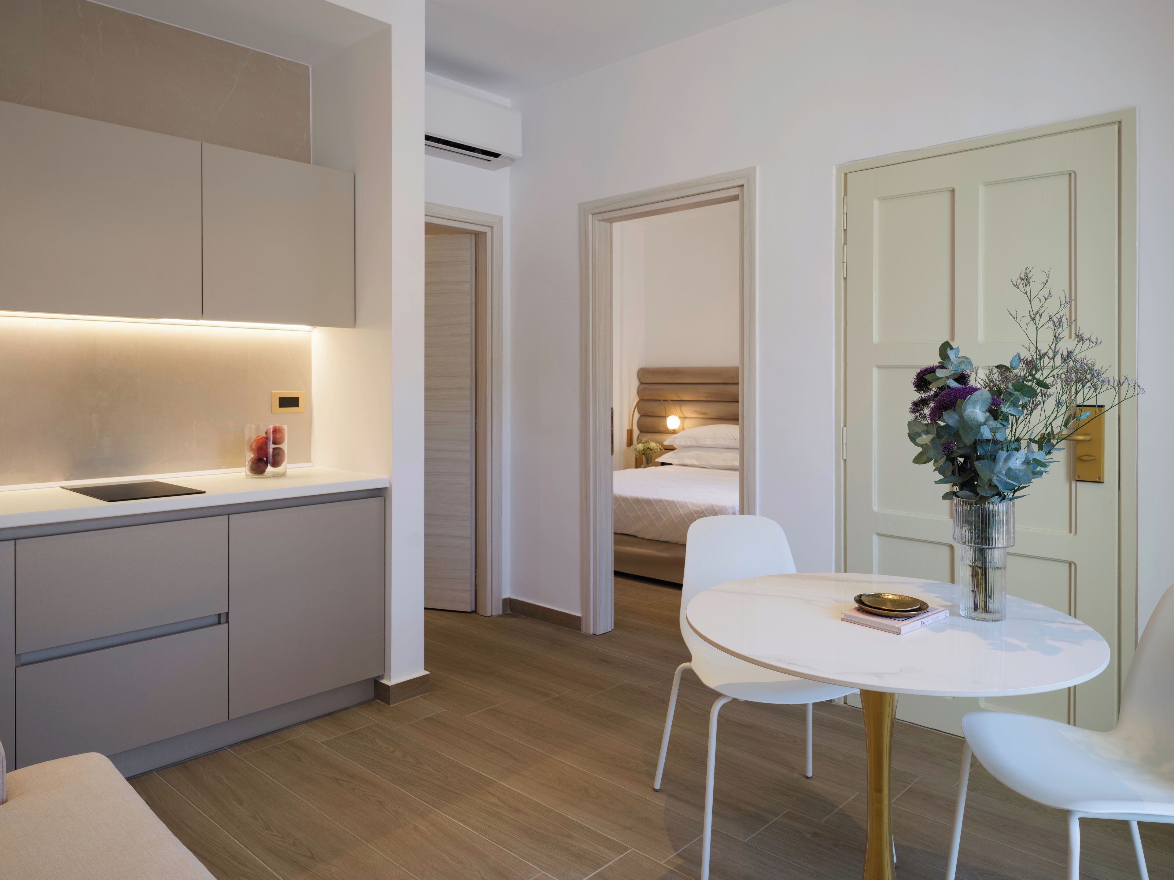 A modern hotel room with a kitchen area, a dining table with chairs, and a bedroom visible through an open door.