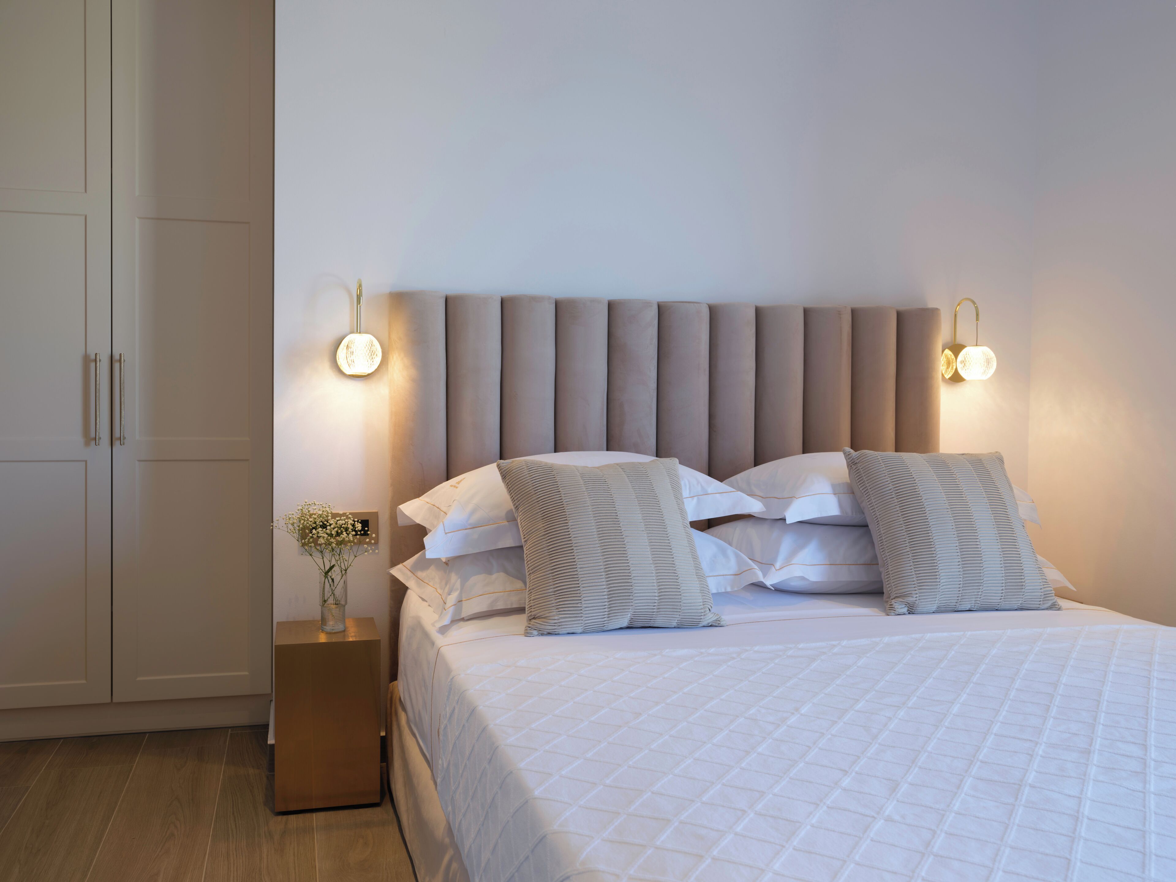 A neatly made bed with a quilted white duvet, two bedside lamps, and a small bedside table with a vase of flowers.