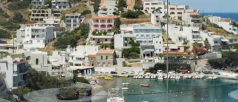 A balcony with a table, a glass of water, and a magazine. The view includes a coastal town with buildings and boats.