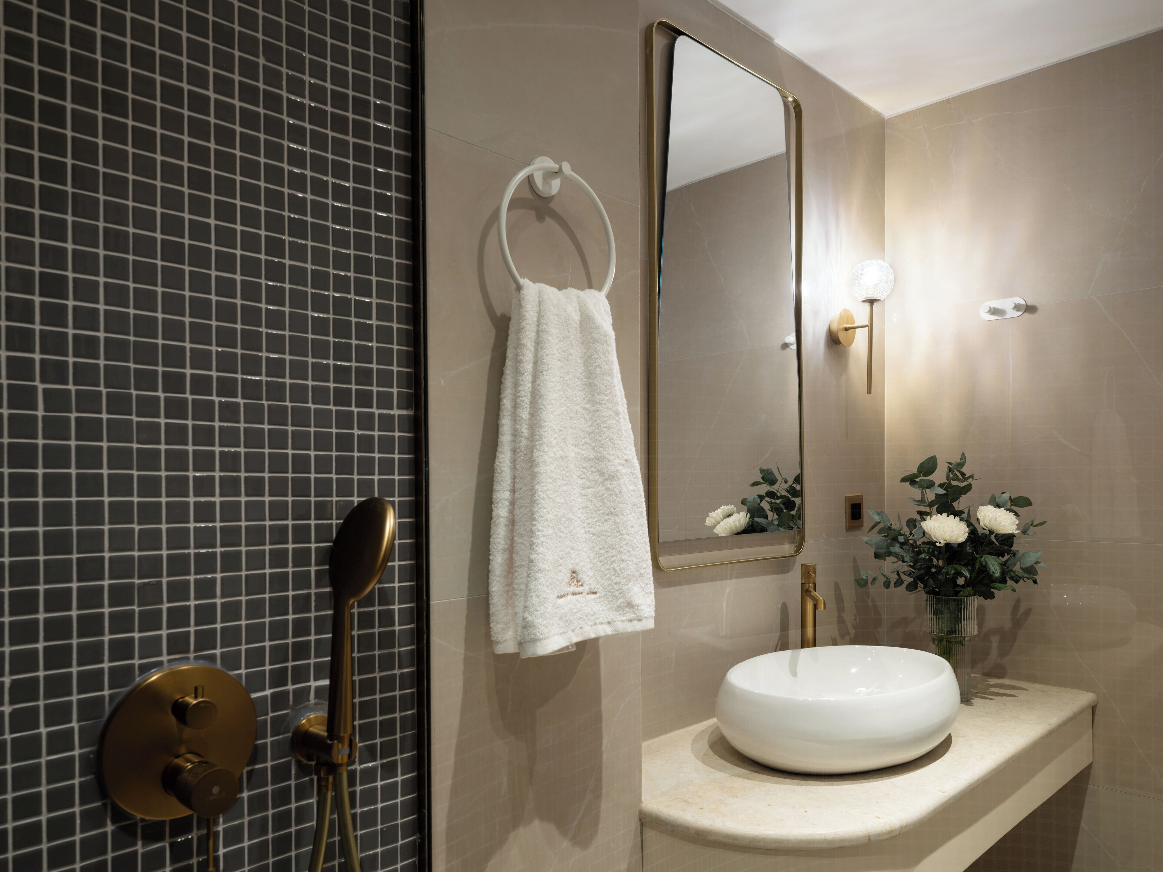 Un baño con pared de azulejos, una toalla blanca colgada en un perchero, un espejo redondo, un grifo dorado y un lavamanos blanco con un jarrón de flores.