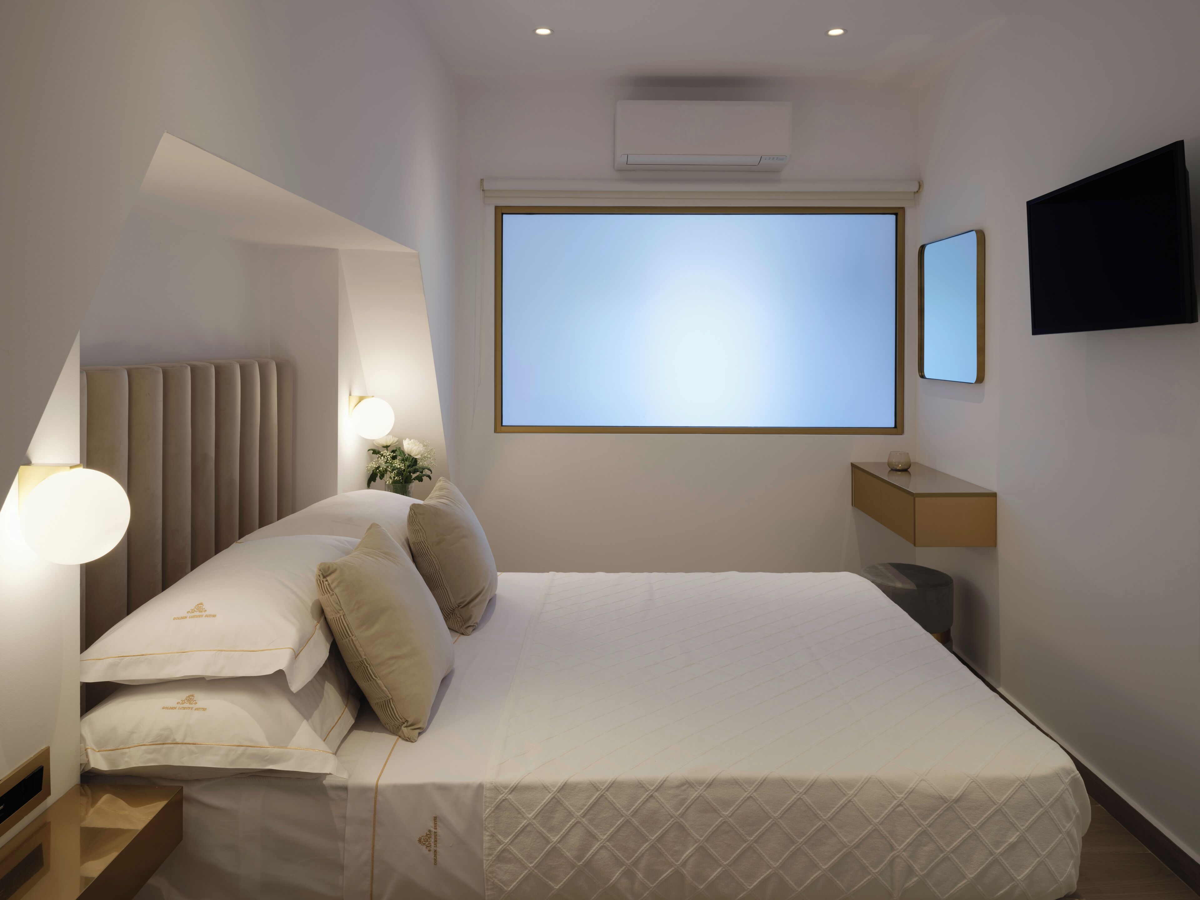 Una habitación de hotel moderna con una cama grande, una mesita de noche, una lámpara y un televisor montado en la pared, y una ventana con vistas.