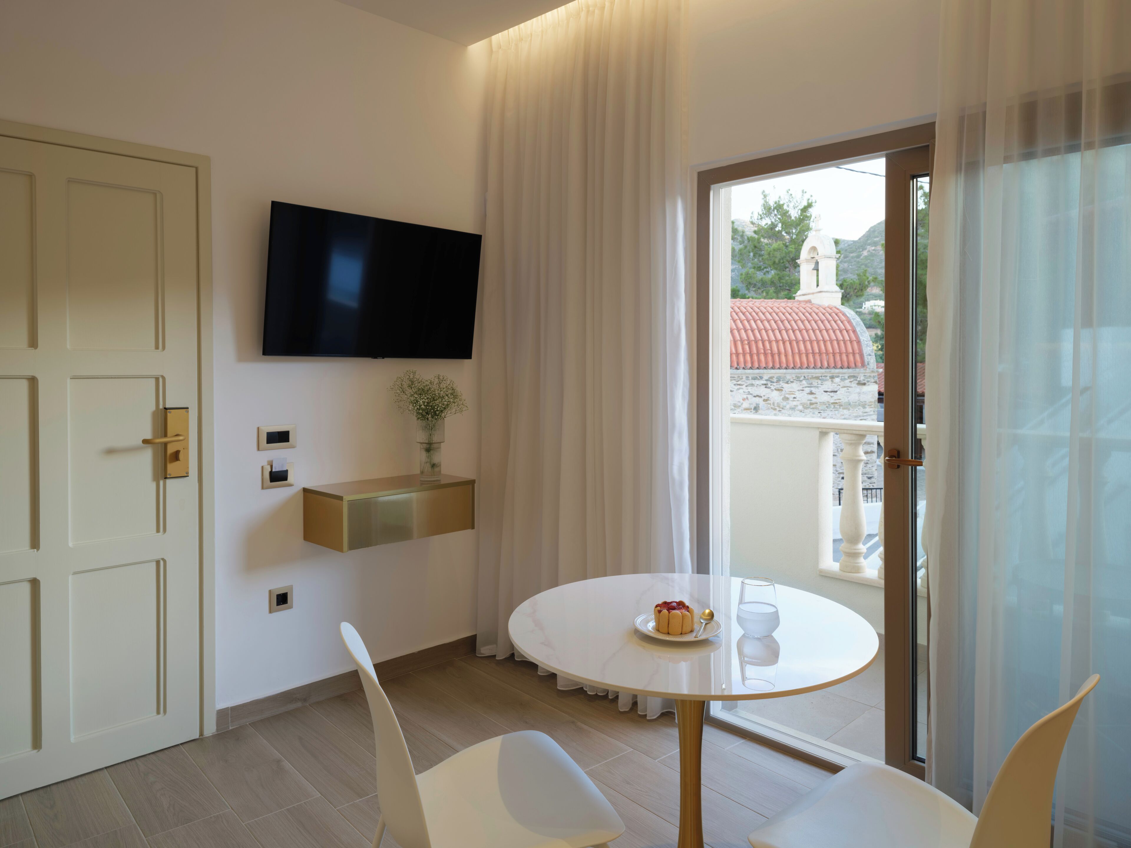 Une chambre d’hôtel moderne équipée d’un téléviseur à écran plat fixé au mur, d’une petite table avec un verre d’eau et une assiette de nourriture, de chaises blanches et d’une porte-fenêtre coulissante donnant sur un espace extérieur avec vue sur un bâtiment au toit rouge.