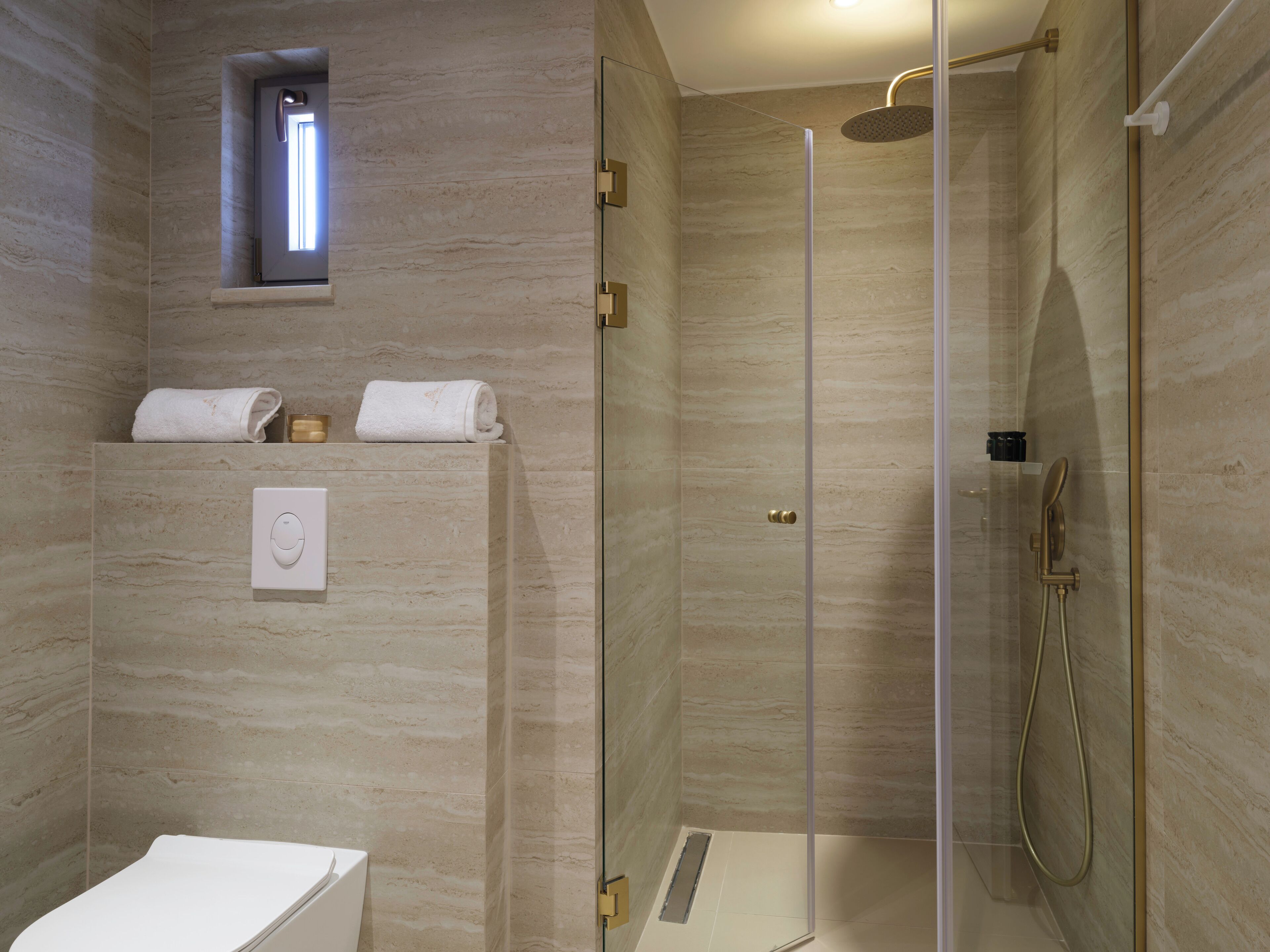 A modern bathroom with a glass-enclosed shower, marble walls, a small window, and a toilet.