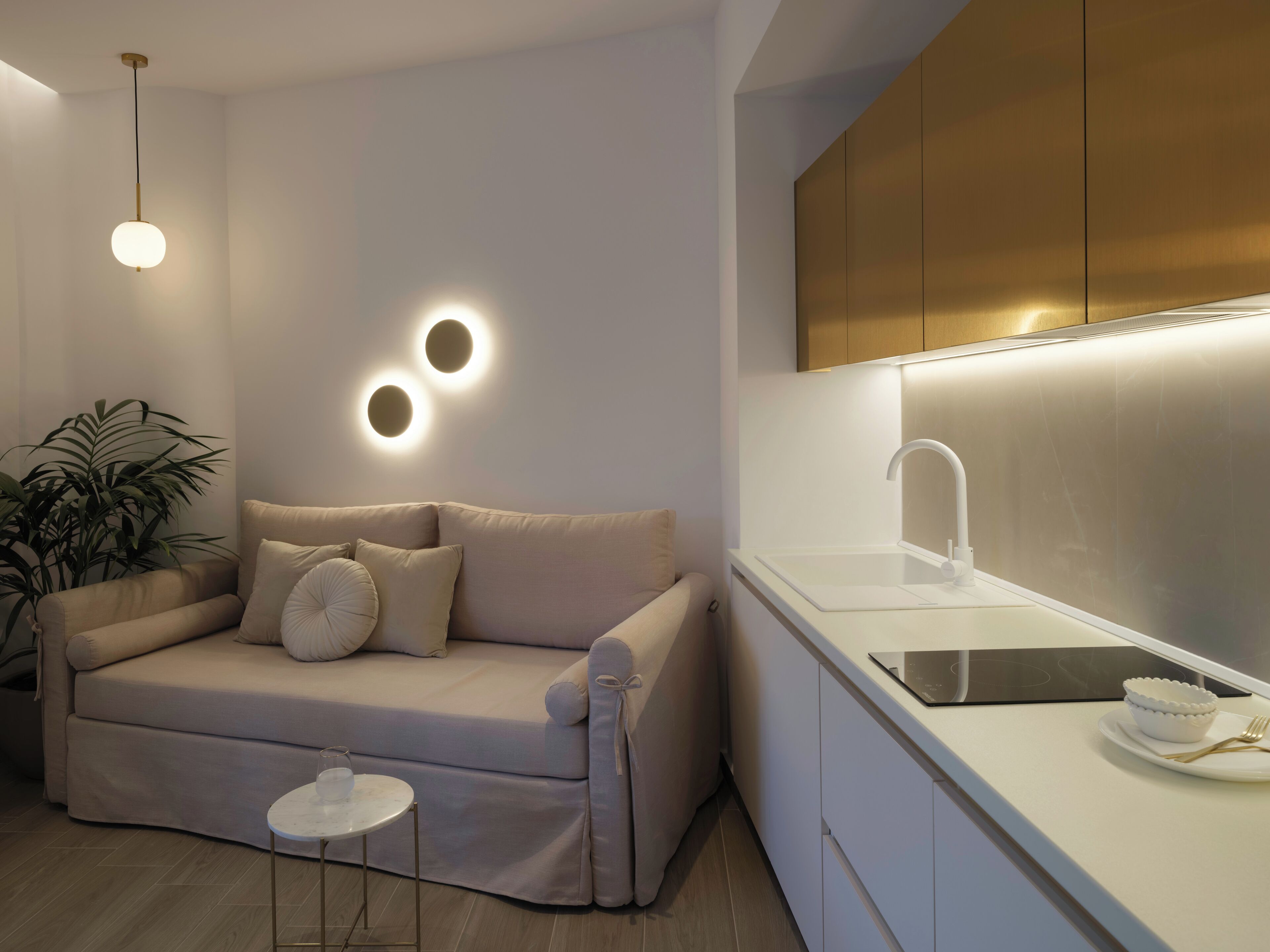 A modern living room with a beige sofa, a small round table, and a kitchen area with a white countertop and sink.