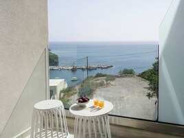 Un balcón con dos sillas blancas y una pequeña mesa con fruta y bebidas, con vista a la costa, barcos y un muelle.