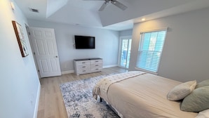 Une chambre à coucher comprenant un lit, un téléviseur, un ventilateur de plafond, une fenêtre avec des stores et une porte.