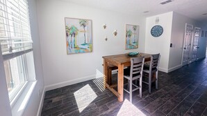 Un coin repas avec une table et des chaises en bois, deux tableaux accrochés au mur, une horloge et une fenêtre avec des stores.