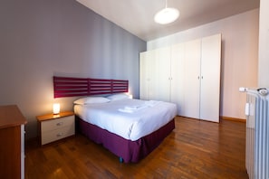 Quarto com piso de madeira, cama com roupa de cama branca, mesa de cabeceira com abajur, guarda-roupa e radiador.