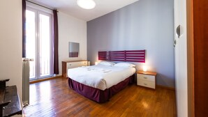 Un dormitorio con piso de madera, una cama con cubrecama blanco y color borgoña, una mesita de noche con lámpara, y una ventana con cortinas.