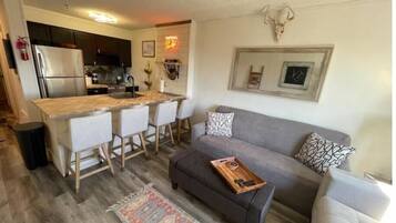 A compact living space with a kitchenette, a gray sofa, a patterned rug, and a wall-mounted skull decoration.
