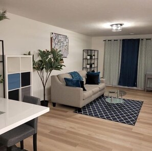 Un salon moderne avec un canapé, une table à manger et une table basse. On y trouve également une plante, une bibliothèque et un tableau accroché au mur.