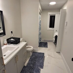 Une petite salle de bain avec un lavabo blanc, des toilettes et une petite fenêtre. Il y a deux tapis sombres sur le sol et un panier sur le comptoir.