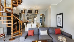 Un salón moderno con un sofá gris, una escalera de madera y una cocina al fondo.