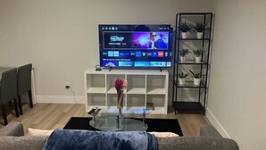 A living room with a flat-screen TV on a stand, a glass coffee table, a gray sofa, a small white shelf with a vase, and a black shelving unit with plants. - A Home away from Home
 (Dieppe)