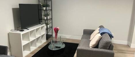 A modern living room with a grey sofa, a flat-screen TV on a white stand, a glass coffee table with a vase of flowers, and a black rug.