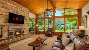 A living room with a fireplace, leather furniture, a television, and large windows with a view of trees. - Stargaze | Views | Hot Tub | Dogs | BBQ | Firepit (Leavenworth)