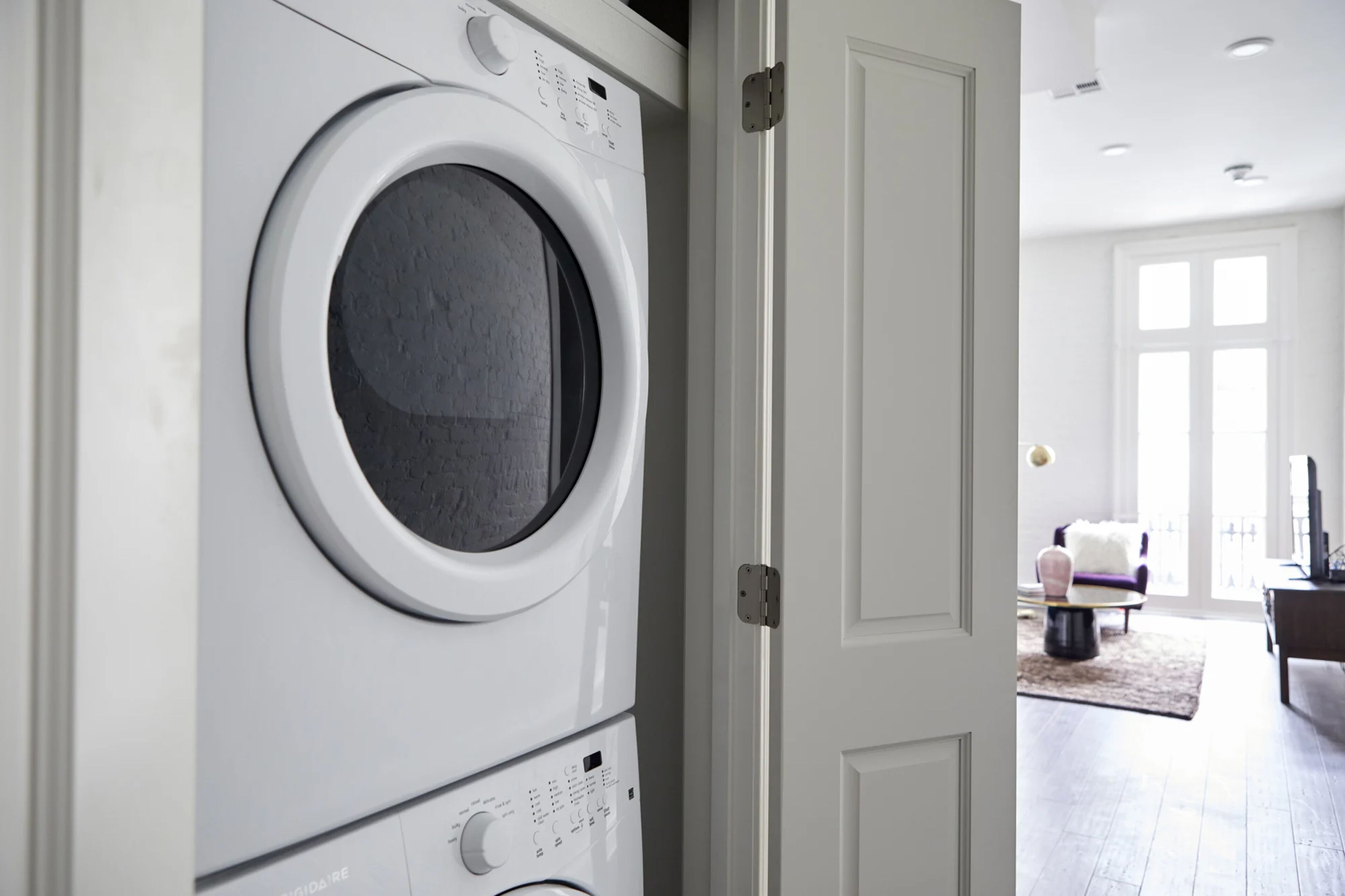 A modern laundry room with stacked washers and dryers, a white door, and a view into a living area with a sofa and a coffee table.