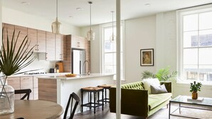 Une cuisine moderne avec des meubles en bois, un îlot central et des luminaires suspendus. On y trouve un canapé vert et une table basse dans un salon doté de grandes fenêtres.