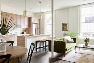 Una cocina moderna con armarios de madera, una isla central e iluminación colgante. Hay un sofá verde y una mesa de centro en un área de estar con ventanas amplias.