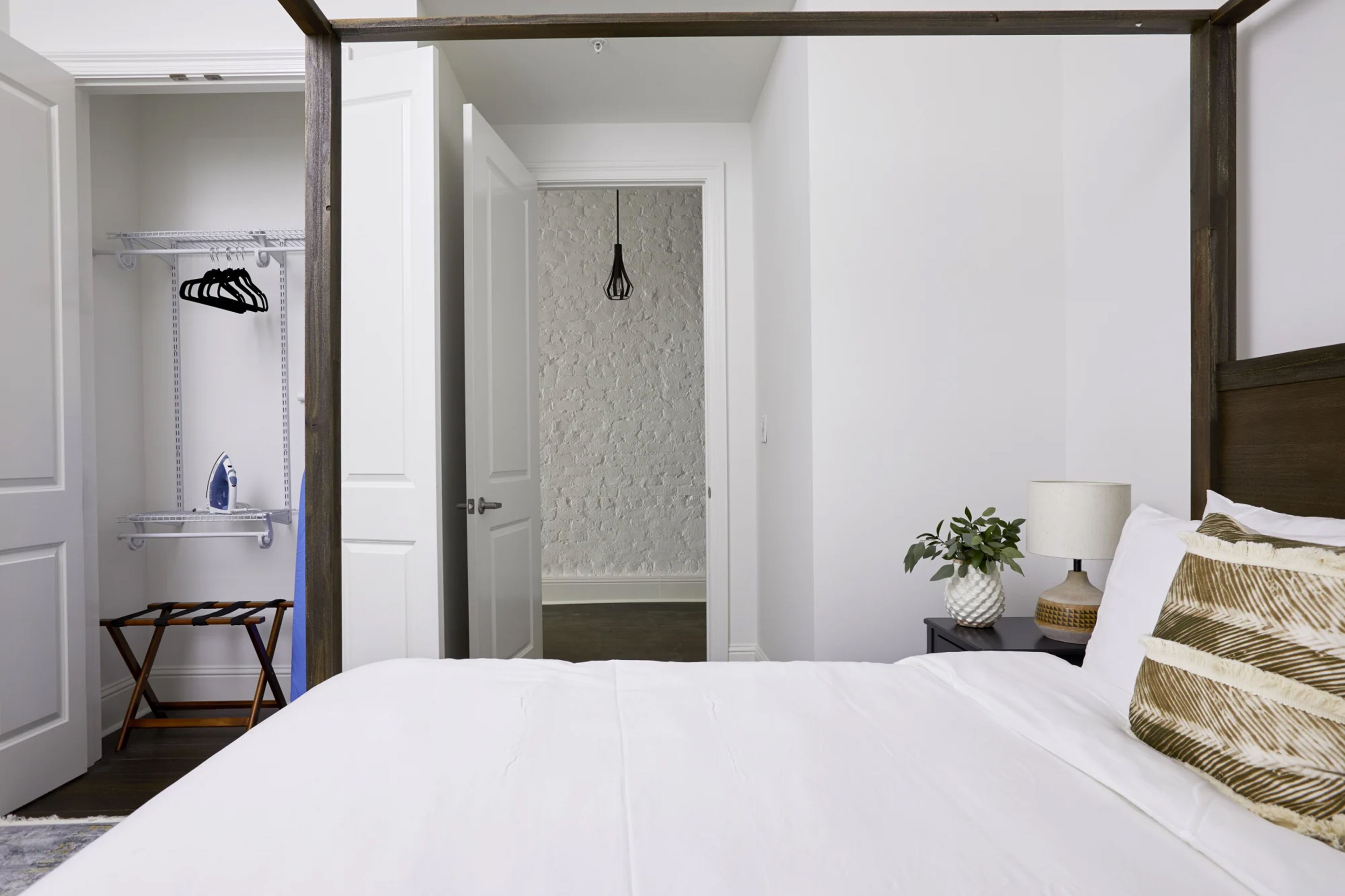 A bedroom with a large bed, a nightstand with a lamp and a plant, a closet with hanging clothes, and a bathroom door.