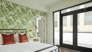 Une chambre à coucher avec un lit, une table de chevet, une plante en pot et une grande fenêtre donnant sur la rue.