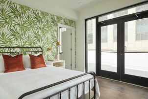 Un dormitorio con cama, mesita de noche, planta en maceta y ventana grande con vista a la calle.