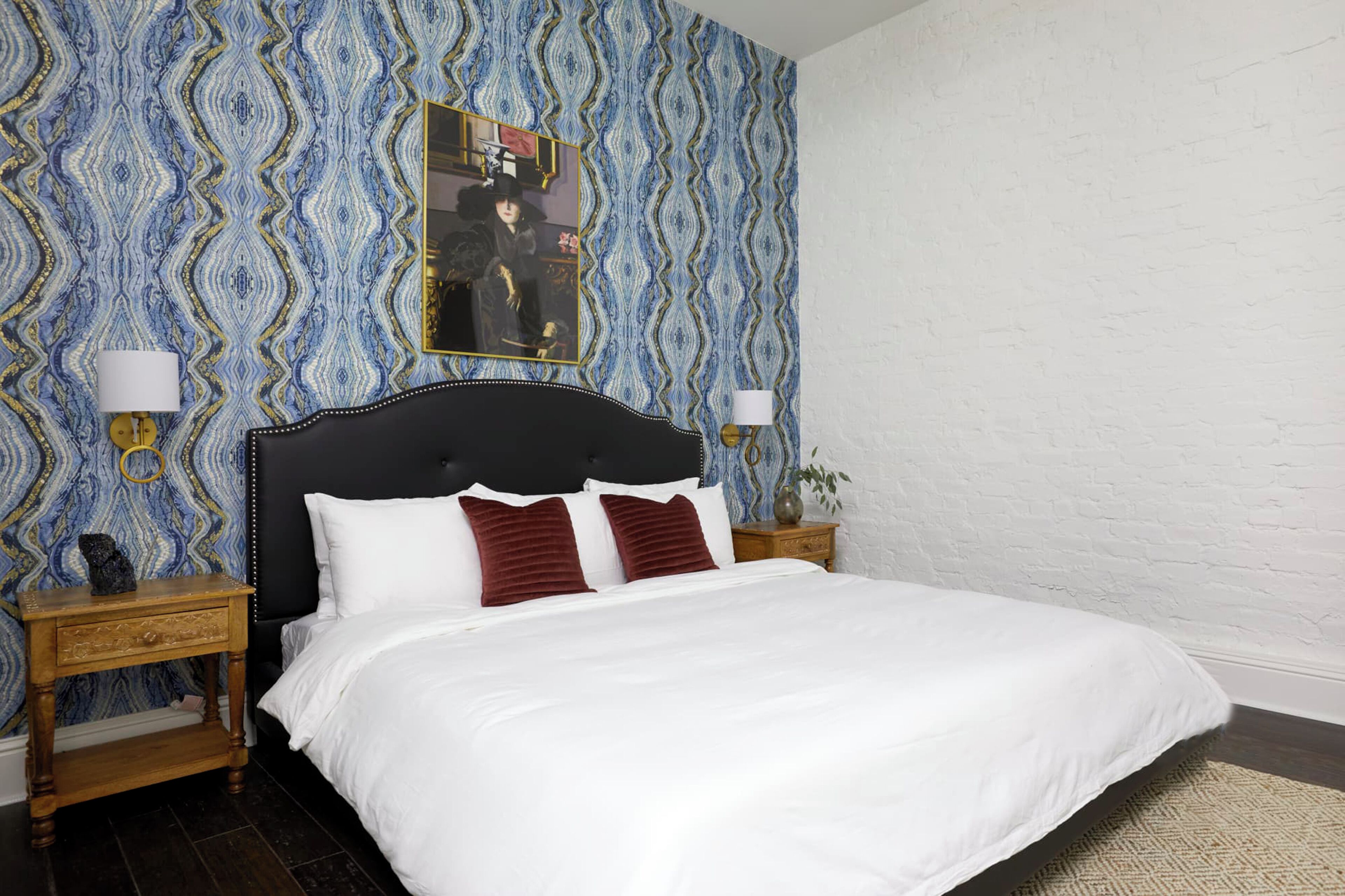 Una cama bien tendida con almohadas blancas y rojas, un cabecero negro y dos mesitas de noche con lámparas. La habitación tiene papel tapiz estampado y una pared de ladrillo blanco.
