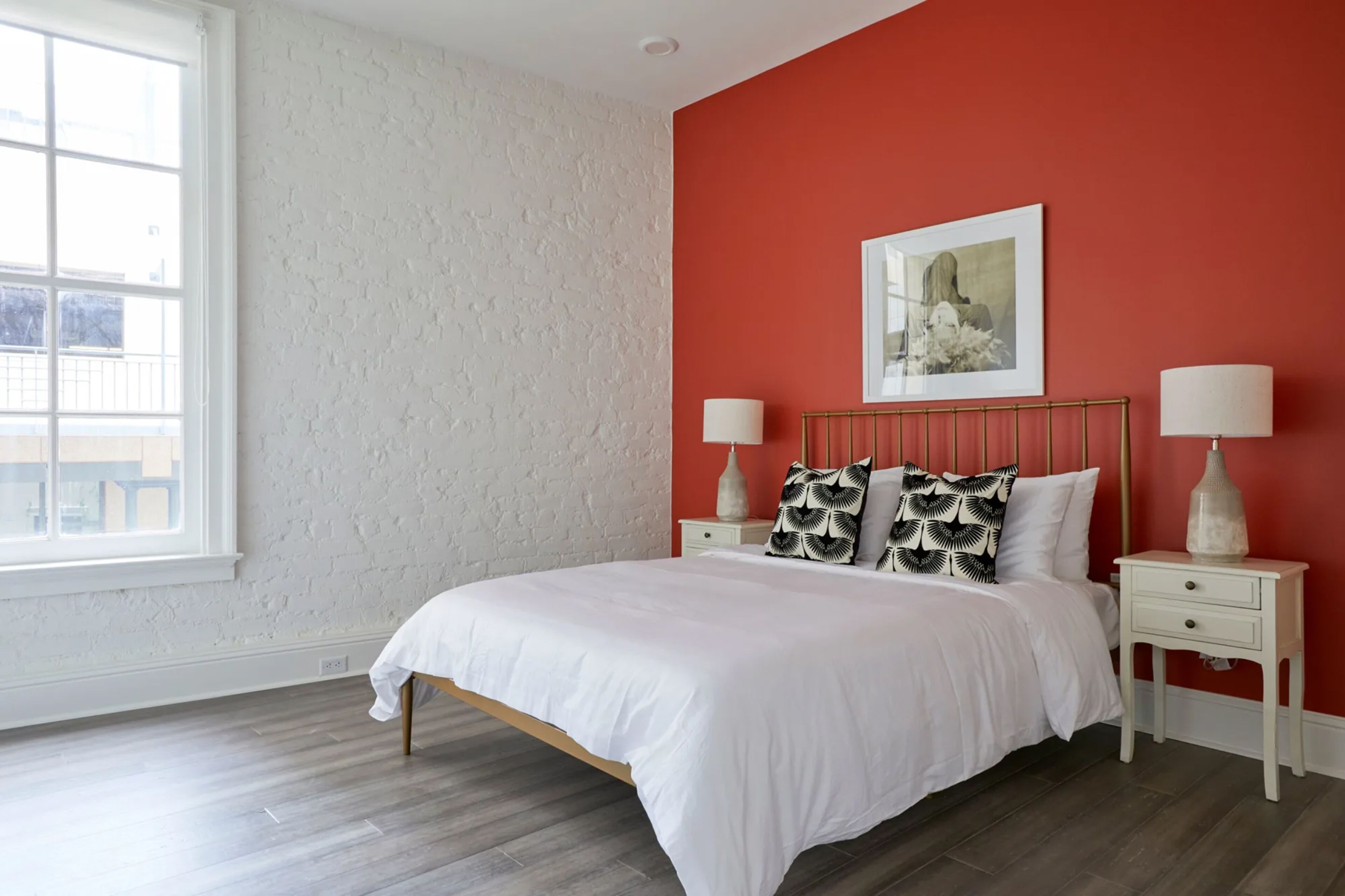 A bedroom with a large window, a bed with white bedding, two bedside tables with lamps, and a red accent wall.