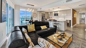 A modern living room with a black leather sofa, a glass coffee table, and a city view through large windows. - Luxury Waterfront Suite with Great Views (Hallandale Beach)
