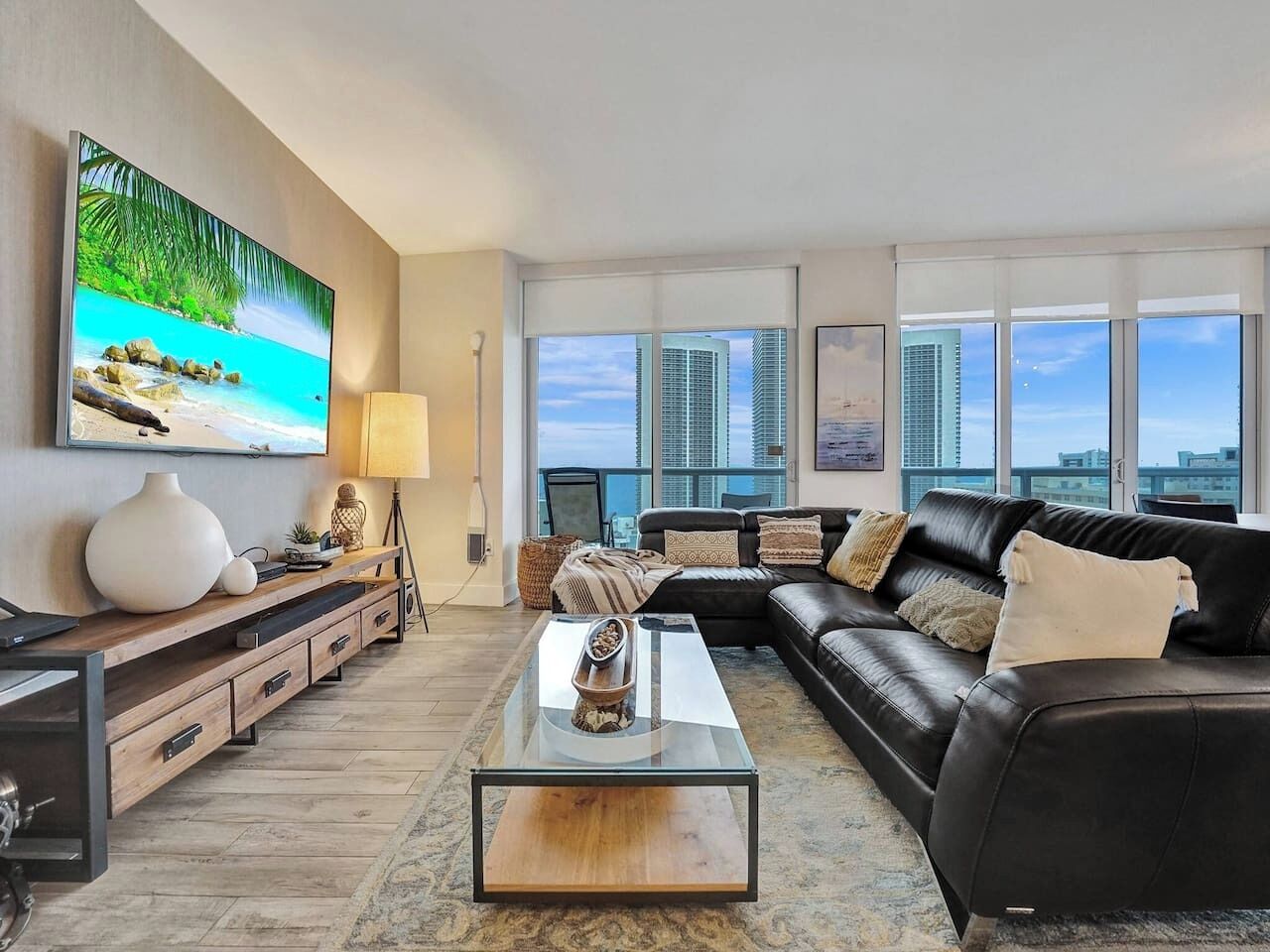 A modern living room with a black leather sofa, a glass coffee table, and a large TV mounted on the wall.