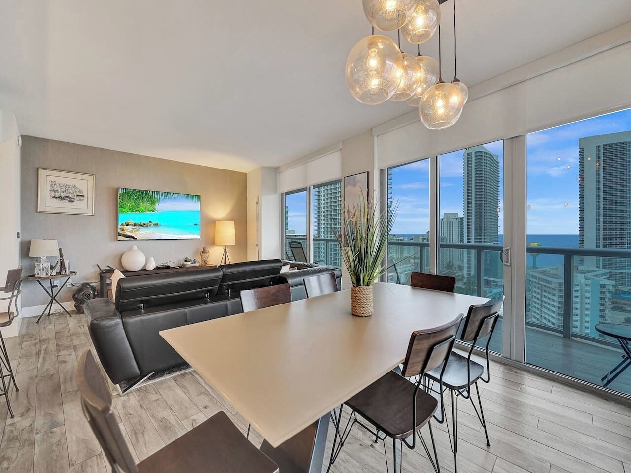 A modern living room with a large dining table, black leather sofa, and a city view through large windows.