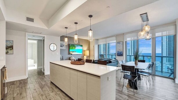 A modern kitchen with a large island, wooden flooring, pendant lights, and a dining area with a view of the city. - Luxury Waterfront Suite with Great Views (Hallandale Beach)