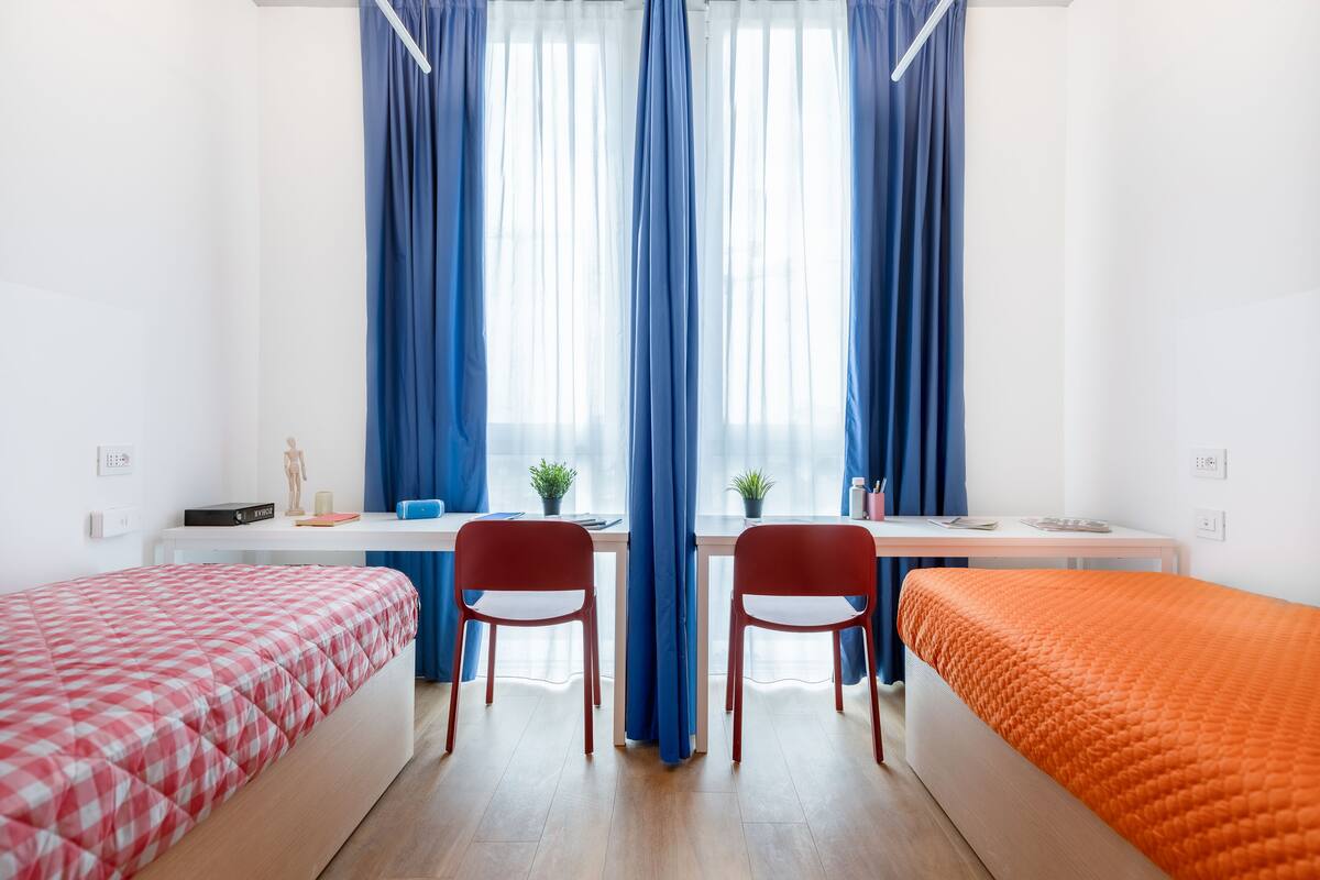 A room with two beds, a desk, chairs, and a window with blue curtains.