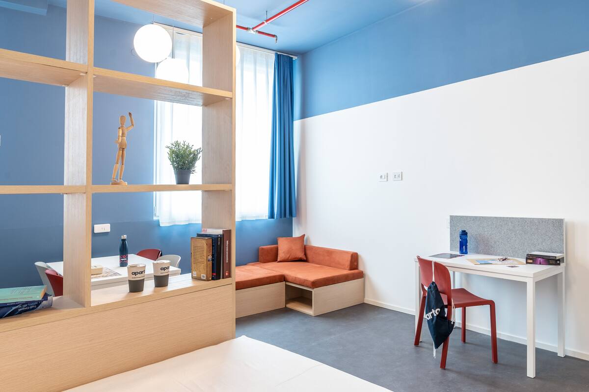 A modern room with a white desk, orange seating, a bookshelf, and a window with blue curtains.