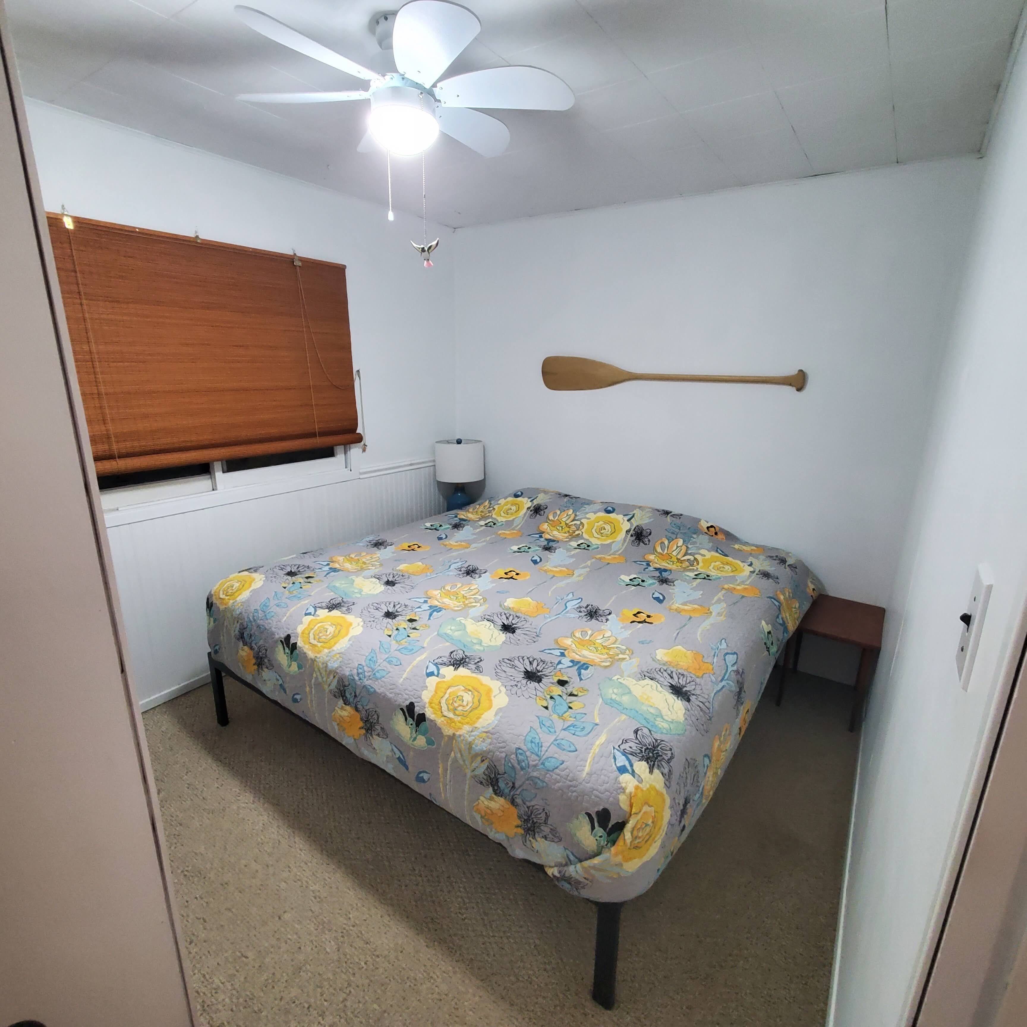 A bedroom with a bed, a ceiling fan, a wooden paddle, a window with blinds, and a bedside lamp.
