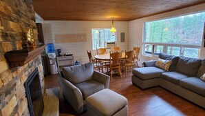 A cozy cabin interior with a stone fireplace, wooden dining table, and a large window offering a view of the outdoors.