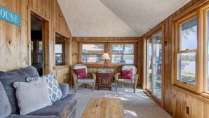 A cozy cabin interior with a wooden exterior, a gray sofa, two wicker chairs, a small table with a lamp, and large windows. - Charming 2-bedroom Hayward Cabin on Lac Courte Oreilles (Hayward)