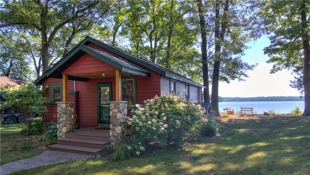A red cabin with a stone foundation, a porch, and a view of a lake. - Charming 2-bedroom Hayward Cabin on Lac Courte Oreilles (Hayward)