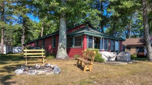 A red cabin with a green roof, a wooden bench, and a stone fire pit in a wooded area. - Charming 2-bedroom Hayward Cabin on Lac Courte Oreilles (Hayward)