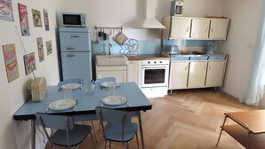 Cozinha compacta com área de refeições, com mesa azul, cadeiras e eletrodomésticos antigos.
