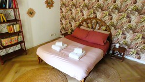 Uma cama bem arrumada com roupa de cama rosa e duas toalhas, uma estante de madeira cheia de livros e um papel de parede floral.