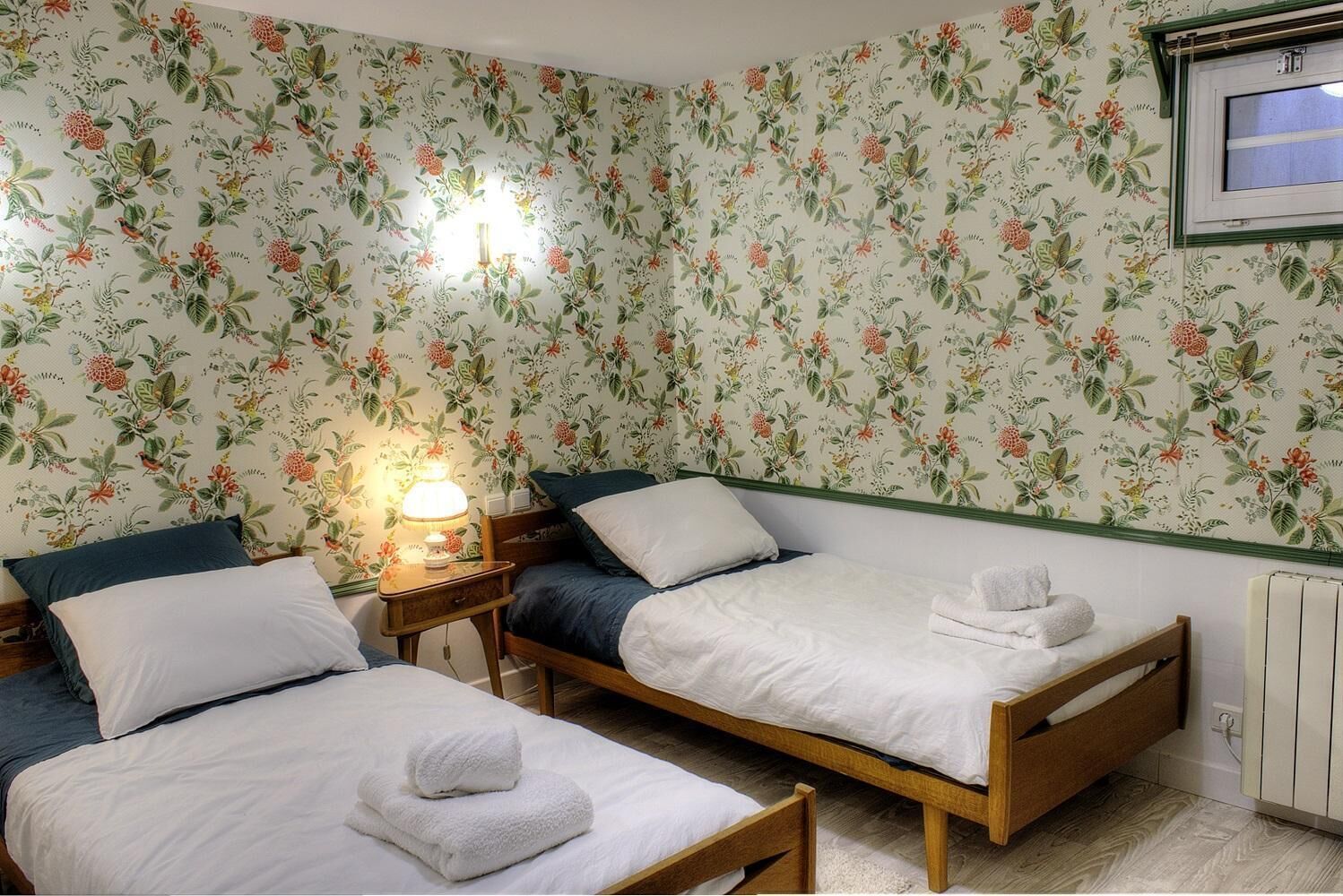 Una habitación con dos camas individuales, una mesita de noche con lámpara, papel tapiz floral y una ventana con vistas.