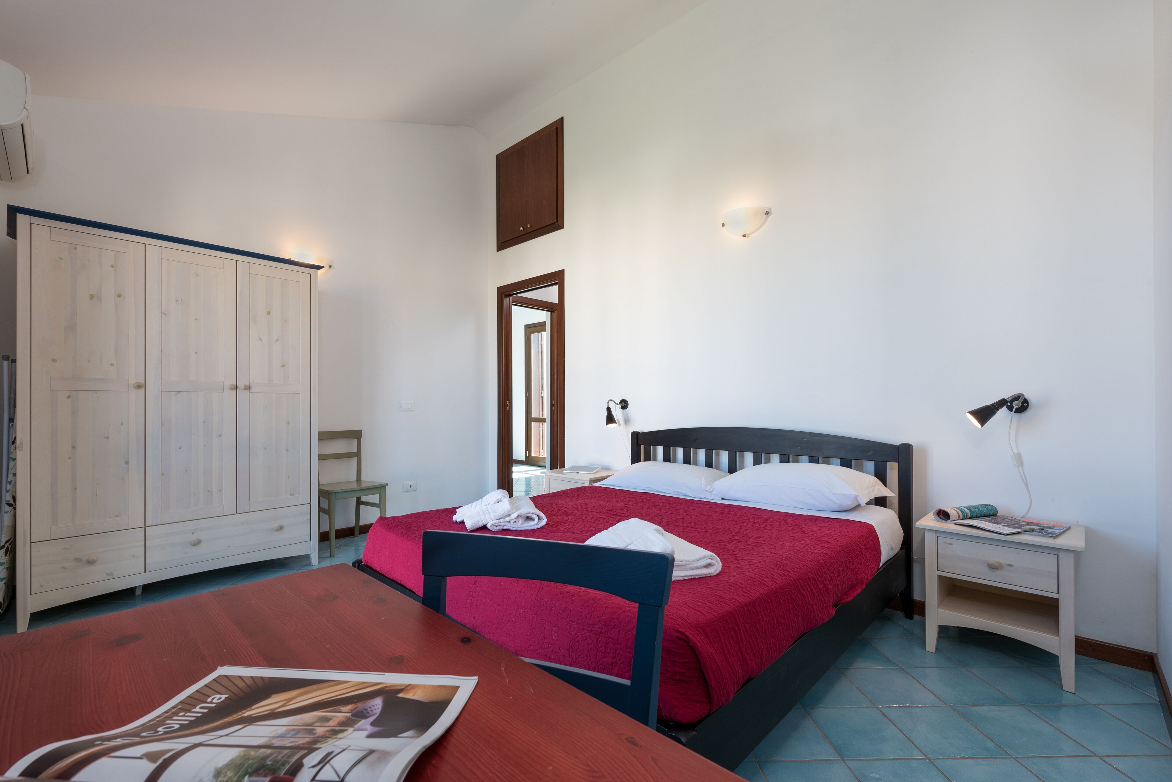 A bedroom with a bed, a nightstand, a wooden wardrobe, and a table with a magazine.