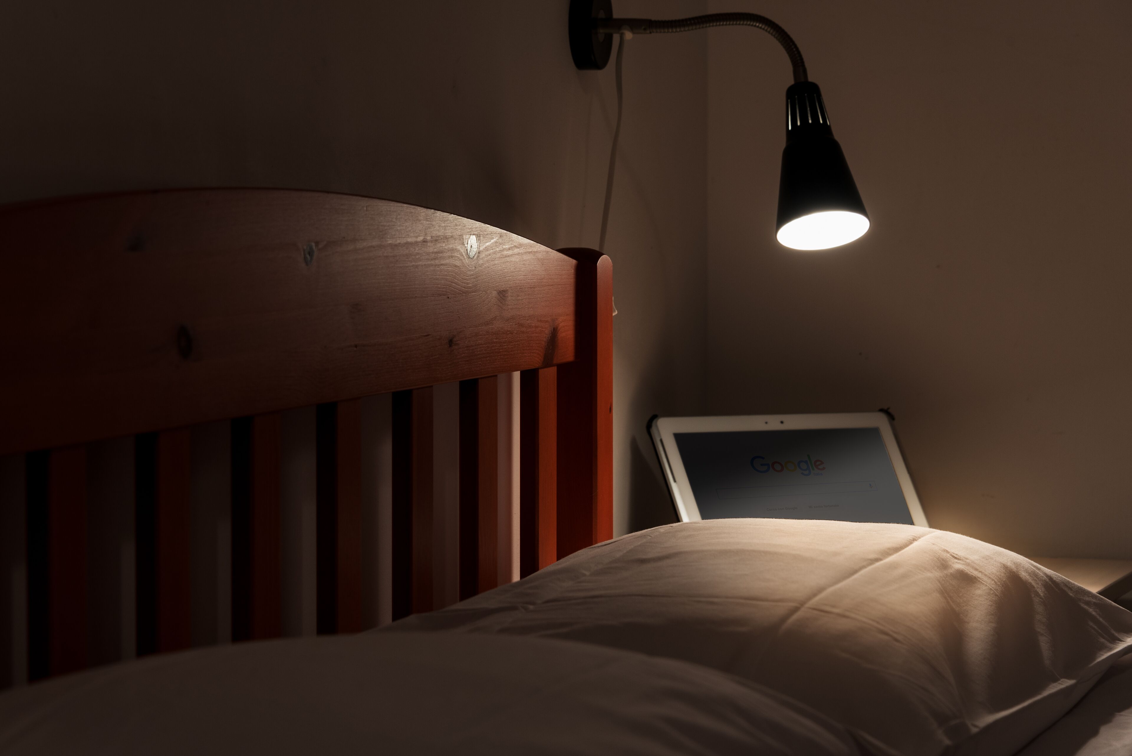 Ein Bett mit Holz-Kopfteil, einer Nachttischlampe und einem Tablet, das die Google-Startseite anzeigt.